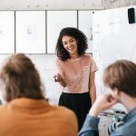 A woman in front of a whiteboard talking to a class.