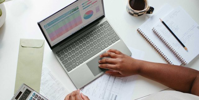 A person using a laptop. On the screen are reports. On the desk a cup of coffee