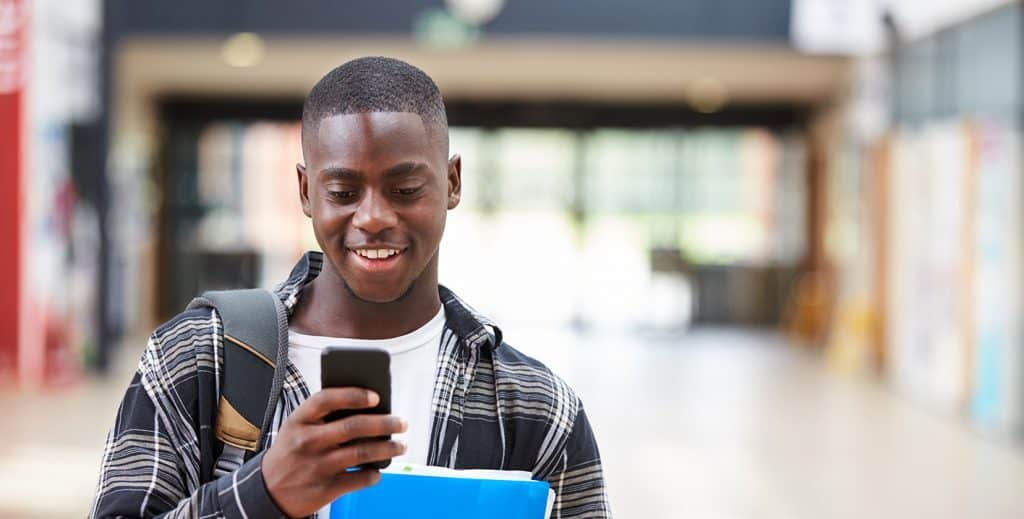 A smiling student looking at his phone while walking down a corridor holding some folders